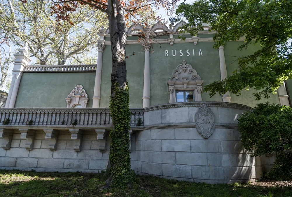 Venedik Bienali Rusya Pavyonu'nun RUSSIA yazısının, 1914 tarihli süslü armanın, zarif balustratın ve ön plandaki sarmaşık kaplı ağacın görüldüğü, yeşilimsi renkli dış cephesi ve doğal çevresiyle bütünleşmiş görkemli bir görünümü.