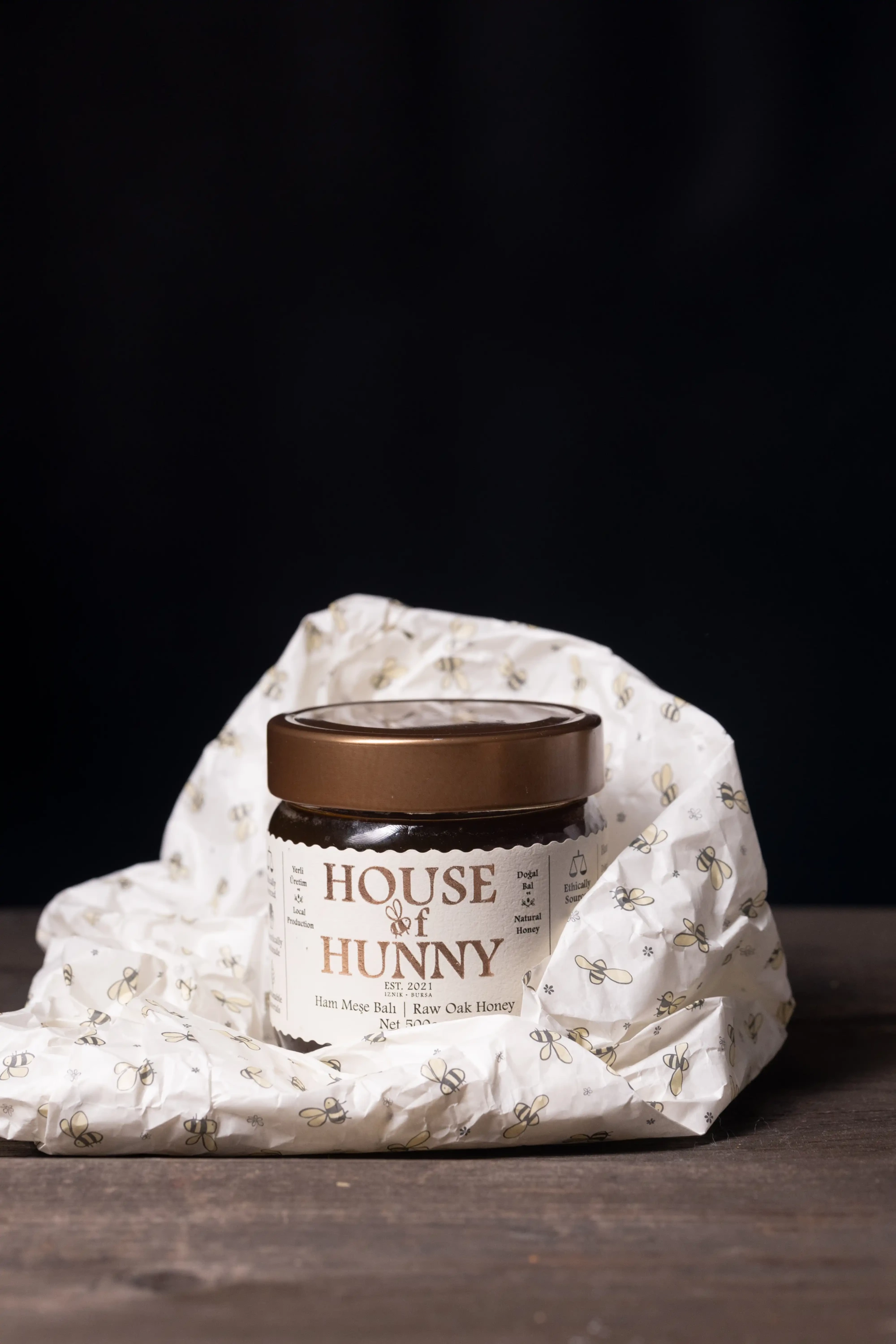 Koyu bronz kapaklı, House of Hunny markalı, yerli üretim doğal ham meşe balı içeren cam kavanoz, arı figürleriyle süslenmiş buruşuk beyaz ambalaj kağıdının üzerinde koyu kahverengi ahşap bir yüzeyde, siyah bir arka planın önünde fotoğrafın merkezinde yer alıyor.