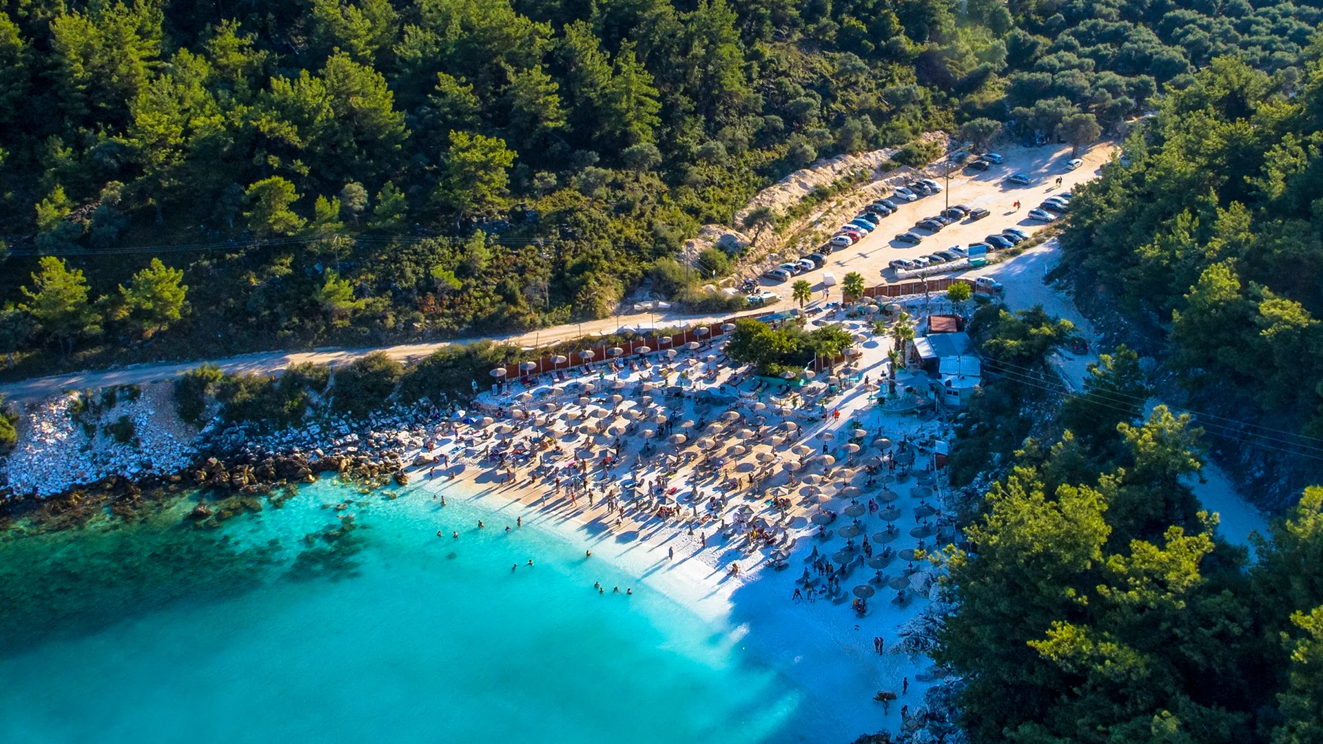 Havadan çekilmiş bu fotoğrafta, turkuaz renkli berrak sularda yüzenler ve kalabalık plajda yan yana dizilmiş şezlonglar ile şemsiyeler dikkat çekiyor; arkasında ise araçlarla dolu bir otopark ve yemyeşil ağaçlarla kaplı tepeler uzanıyor.
