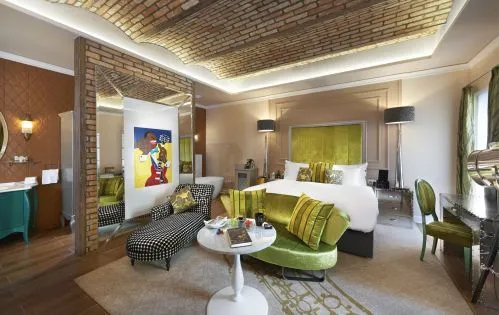 Aynalı duvarlar ve tuğla tavanlı şık bir otel odasında, yeşil kadife bir kanepenin önünde küçük beyaz bir masa, beyaz çarşaflı konforlu bir yatak ve desenli bir dinlenme şezlongu bulunuyor.