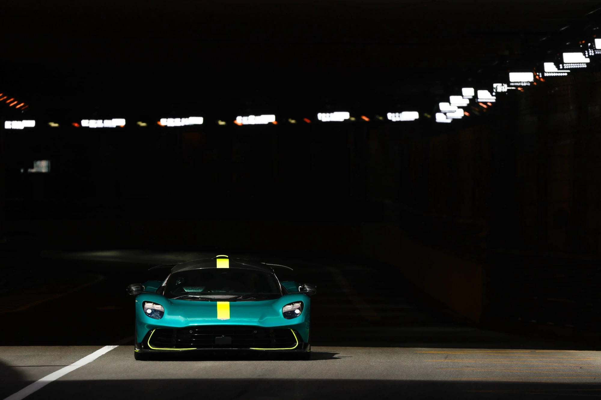 Karanlık bir tünelde ön farları yanık, kaputu ve tavanında sarı bir şerit bulunan çarpıcı teal rengi Aston Martin Valhalla, Formula 1 teknolojisiyle geliştirilen radikal aerodinamik tasarımıyla tüm gücünü sergiliyor.