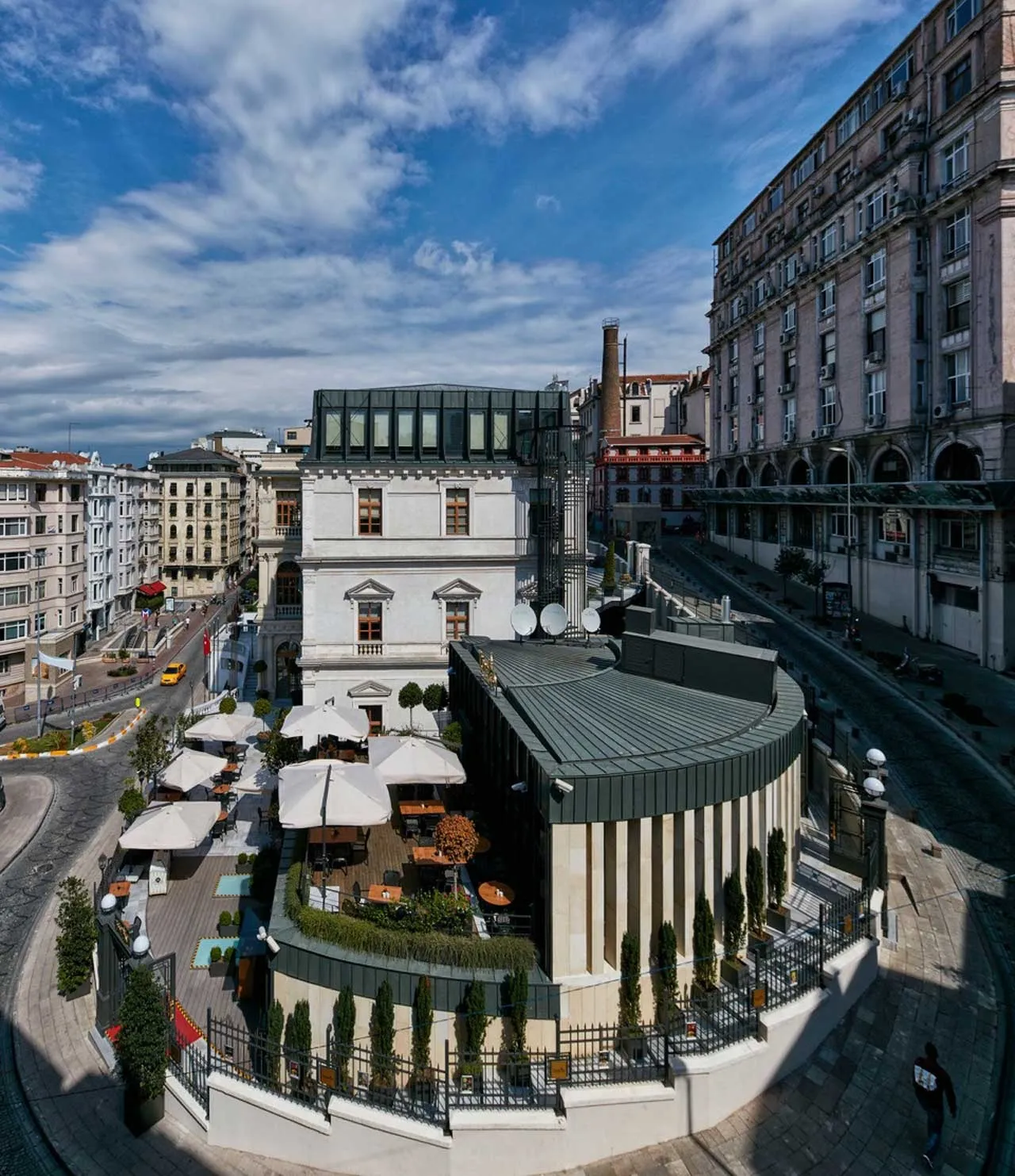 Beyoğlu Belediyesi Binası'nın ve bitişiğindeki kavisli modern yapının, şık beyaz şemsiyelerle donatılmış ahşap teraslı açık hava restoranının kuşbakışı görüntüsü, arka plandaki yoğun şehir dokusu ve masmavi gökyüzüyle birlikte hareketli bir kent manzarasını yansıtıyor.