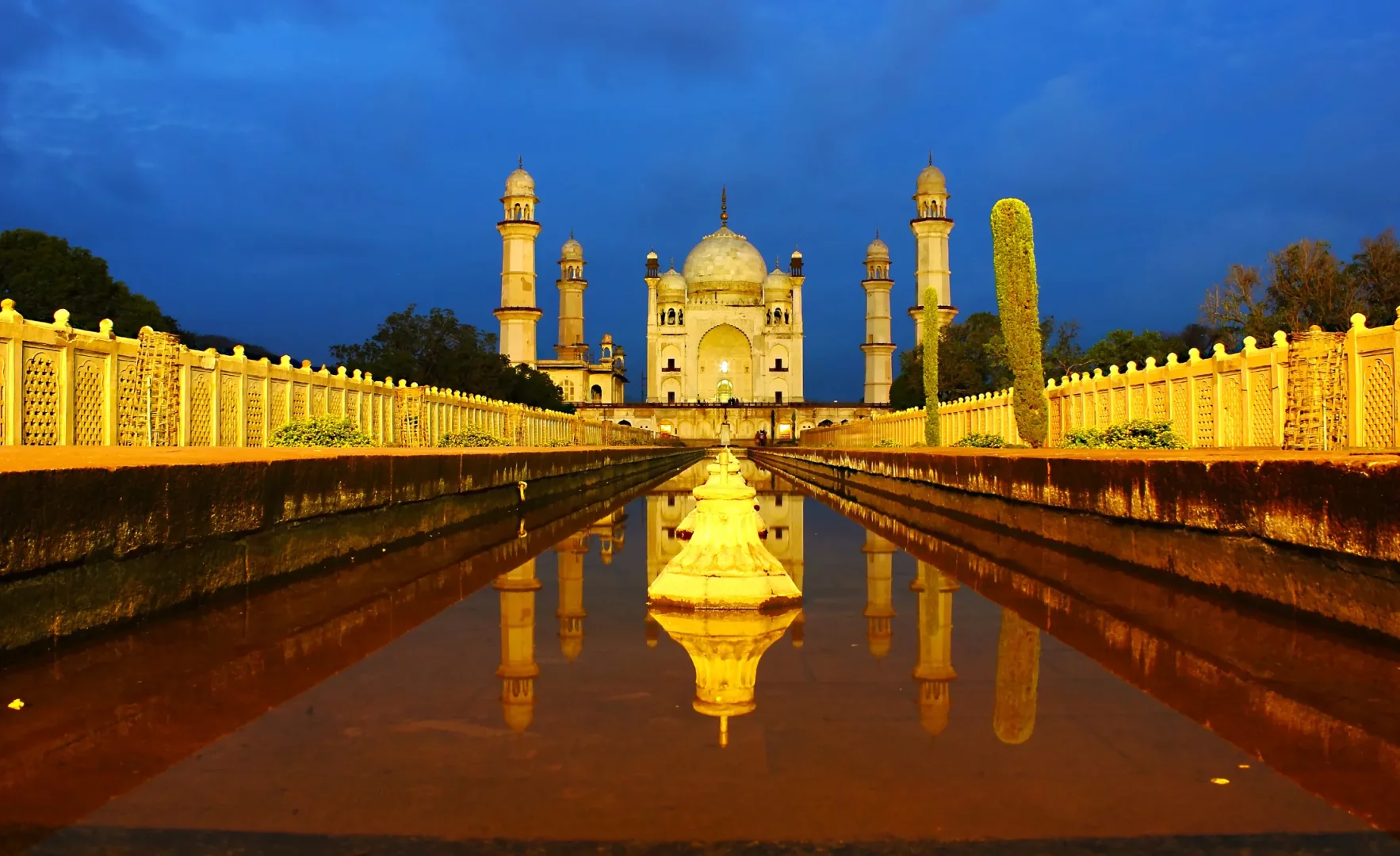 Bibi Ka Maqbara'nın alacakaranlıkta çekilmiş nefes kesici bir görüntüsü, görkemli beyaz mermer mozolesi ve dört uzun minaresi, ön plandaki sakin su kanalında mükemmel bir şekilde yansırken, anıta doğru uzanan sıcak ışıklarla aydınlatılmış dekoratif duvarlar derin mavi bir gökyüzünün altında yer alıyor.
