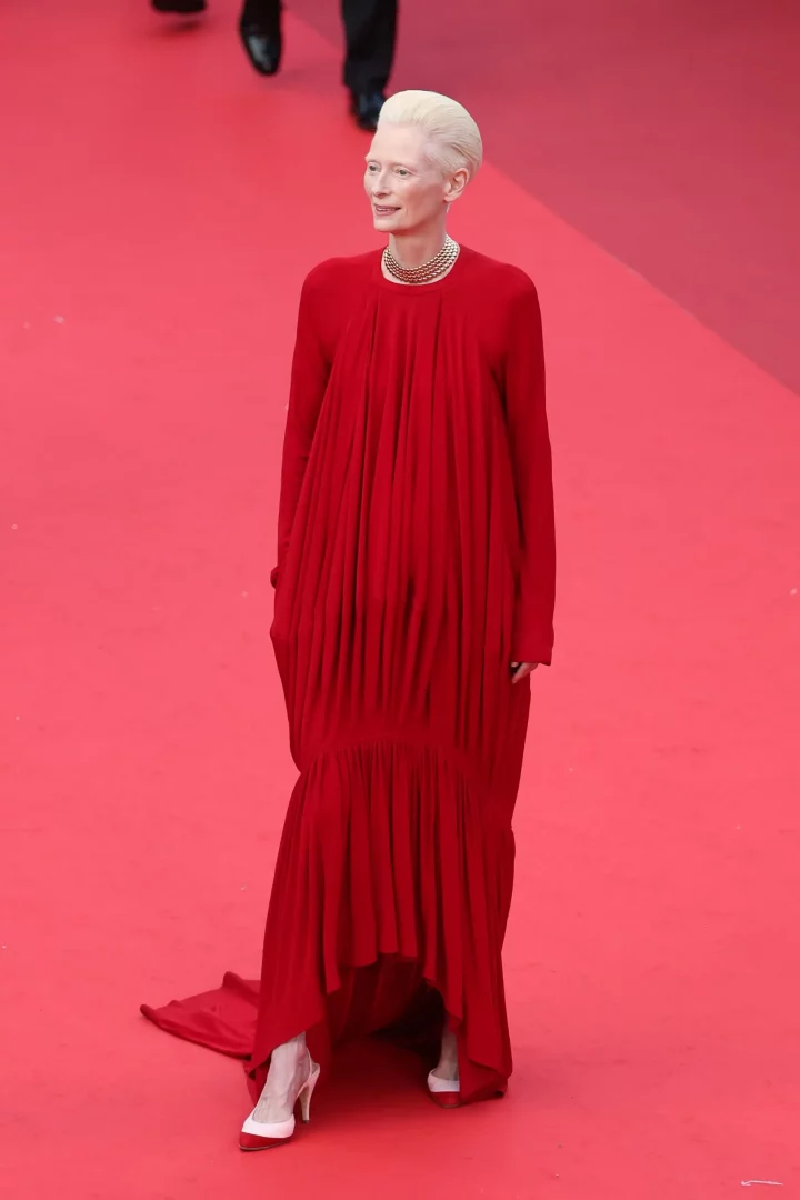 2026 Cannes Film Festivali kırmızı halı görünümlerinden birinde, kısa platin sarısı saçları ve zarif gülümsemesiyle dikkat çeken Tilda Swinton, çok katlı altın renkli bir choker kolye ile tamamladığı, bol kesim ve dökümlü, uzun, parlak kırmızı elbisesi ve beyaz-kırmızı topuklu ayakkabılarıyla şıklığını sergiliyor.