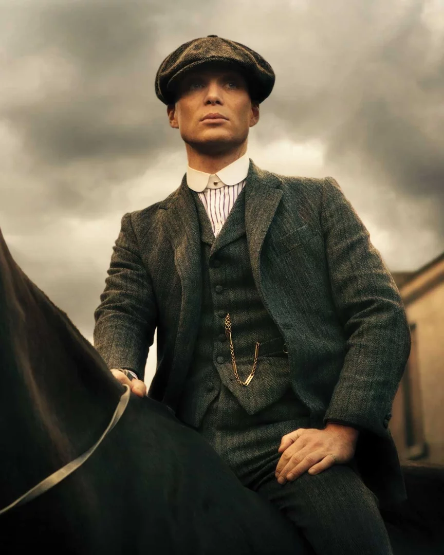 Cillian Murphy, Peaky Blinders dizisindeki kararlı duruşuyla, tweed kasketi ve koyu gri takım elbisesi içinde, yeleğinden sarkan zarif altın cep saati zinciriyle İngiliz modasına özgü bir tarzı bulutlu bir gökyüzü altında at üzerinde sergiliyor.