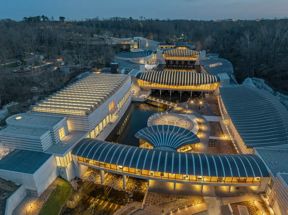 Crystal Bridges Sanat Müzesi'nin havadan çekilmiş alacakaranlık görüntüsünde, ormanlık araziye ustaca yerleşen, kavisli ve çizgili çatılara sahip modern binalar, ışıl ışıl iç mekanları ve aralarından geçen yansımalı su yolları göz kamaştırıcı bir uyum sergiliyor.