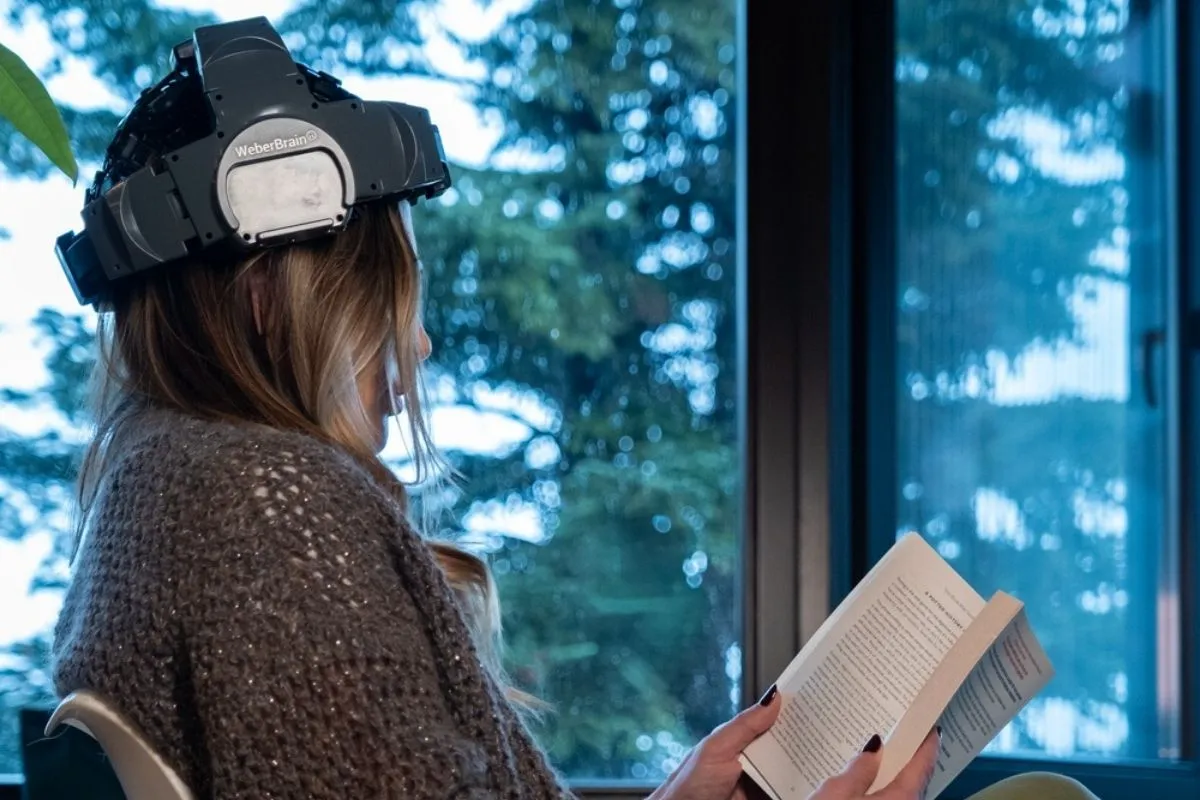 Pencere kenarında oturmuş, başına takılı WeberBrain nörofeedback cihazı ve elinde bir kitapla, uzun ömür ve zihinsel sağlık rehberi kapsamında beyin aktivitesini destekleyerek okuma yapan sarışın bir kadın.