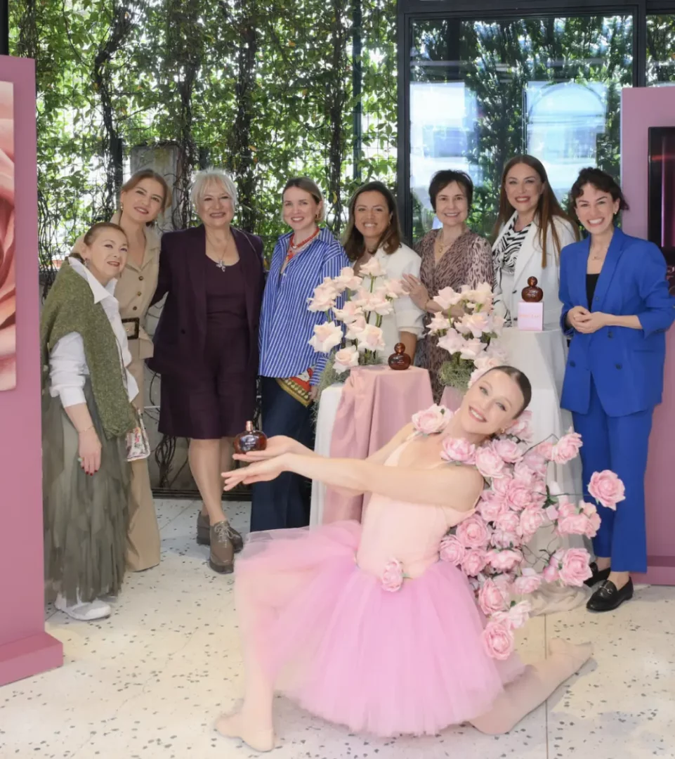Ortada, pembe bir tütü giymiş ve etrafı pembe güllerle sarılmış bir balerin yerde diz çökmüş, elinde küçük bir parfüm şişesi tutarak neşeyle kameraya gülümsüyor. Arkasında, pembe örtülü masaların üzerinde gül aranjmanları ve aynı şişelerle birlikte, çeşitli şık kıyafetler içinde 8 kadın daha gülümseyerek poz veriyor; arka planda yeşil bitkiler ve büyük cam pencerelerle çevrili şık bir etkinlik alanı görülüyor.