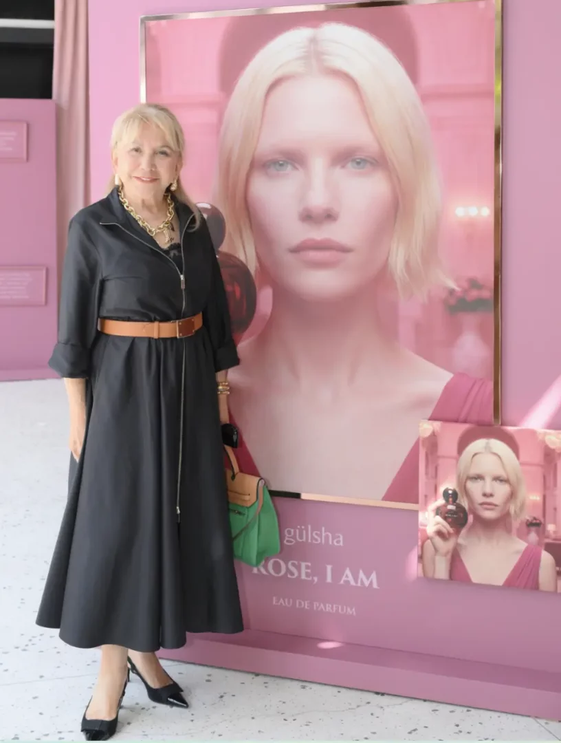 Şık siyah elbisesi, kahverengi kemeri ve gösterişli altın zincir kolyesiyle gülümseyen sarışın bir kadın, büyük pembe gülsha ROSE, I AM EAU DE PARFUM reklam panosunun önünde duruyor; panoda aynı sarışın modelin yakın çekim portresi ve parfüm şişesini tutan bir görüntüsü yer alıyor.