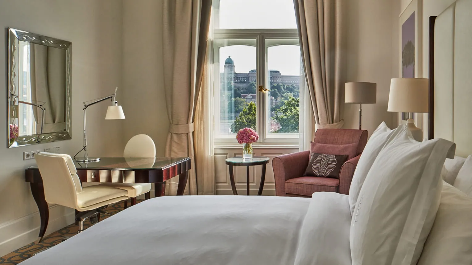 Four Seasons Hotel Gresham Palace'ın şık bir otel odasında, beyaz çarşaflı geniş bir yatak ön plandayken, sol tarafta aynalı bir çalışma alanı ve büyük bir pencereden Budapeşte'nin yemyeşil tepeleri üzerindeki tarihi yapı manzarası eşliğinde konforlu bir koltuk yer alıyor.