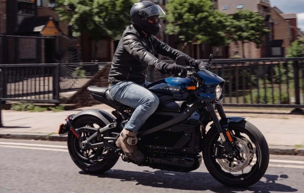Koyu mavi renkli Harley-Davidson LiveWire ONE elektrikli motosikletiyle şehir içinde ilerleyen, siyah kasklı, deri ceketli ve kot pantolonlu bir sürücü, hızla geçerken arka plan hafifçe bulanık görünüyor.