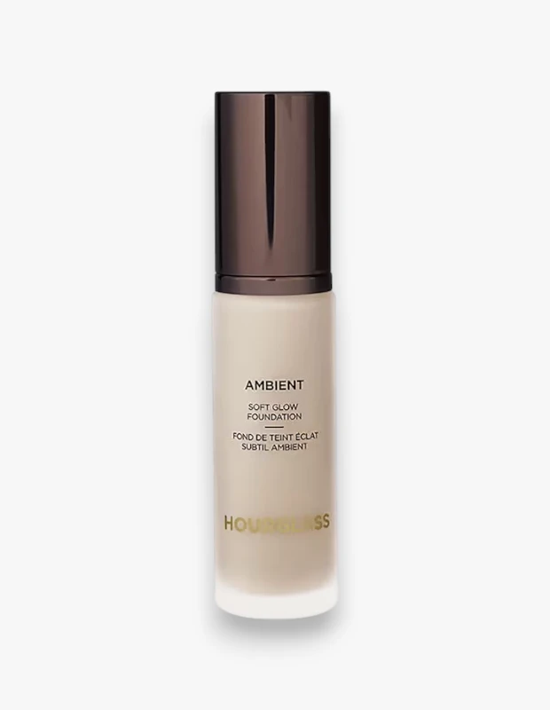 Beyaz bir arka plan üzerinde, alt kısmı buğulu camdan üst kısmı şeffaf olan, açık bej renginde bir Hourglass Ambient Soft Glow Foundation şişesi, parlak koyu bronz kapağı ve üzerinde AMBIENT ile SOFT GLOW FOUNDATION yazılarıyla ürünü belirgin bir şekilde sergiliyor.