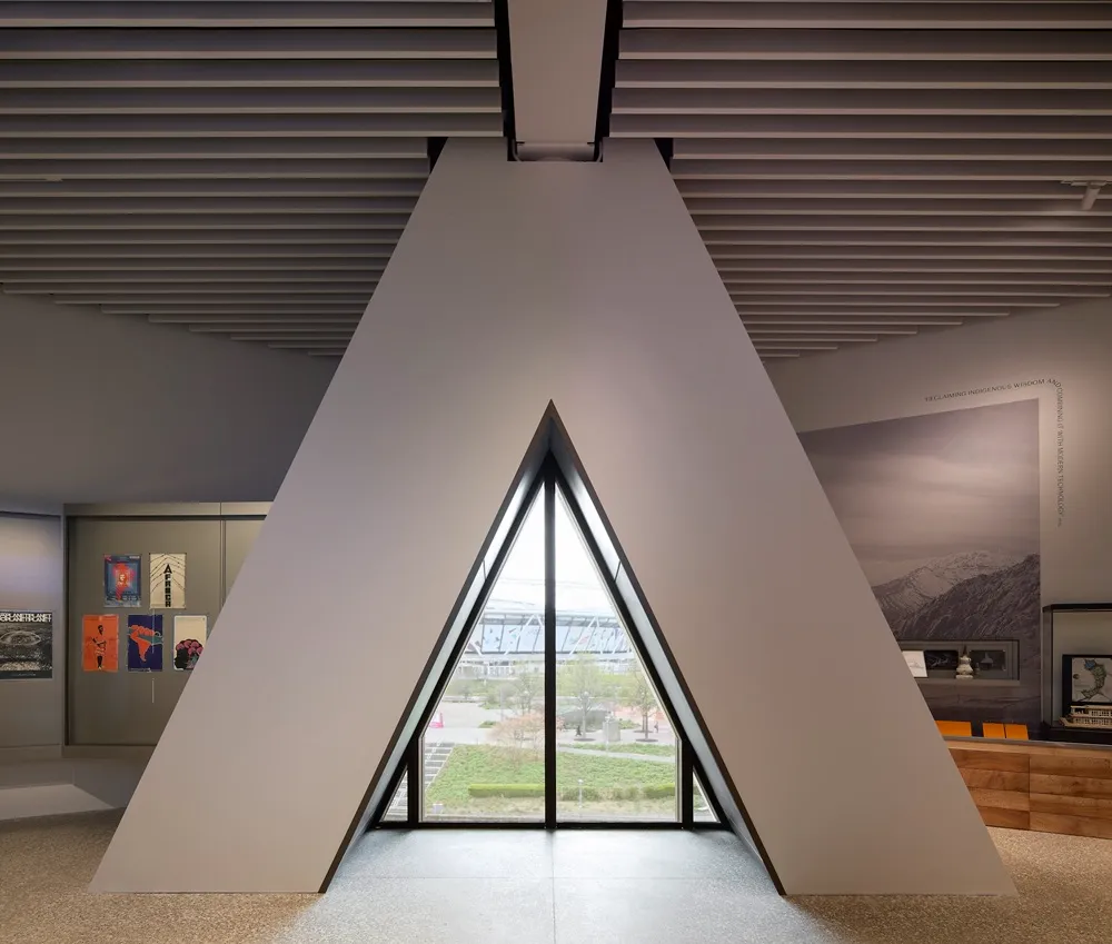 V&A East Müzesi'nin içinde, tavandaki yatay panellerin altında yükselen modern, açısal bir duvar, dışarıdaki yeşil alanı ve uzaktaki stadyumu gösteren üçgen bir pencereye açılırken, sol duvarda renkli posterler ve sağda dağ temalı büyük bir duvar resmi ile sergi alanı görünmektedir.