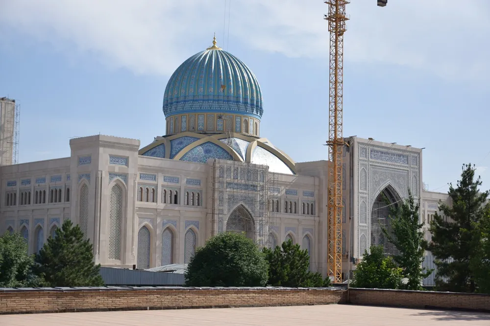 Tuğla duvarlı bir terastan görünen Taşkent İslam Medeniyetleri Müzesi, lacivert ve açık mavi çinilerle bezeli görkemli kubbesi, zarif kemerli pencereleri ve inşaat vinci ile iskeleleriyle devam eden yapım çalışmasını sergileyen bir yapı.