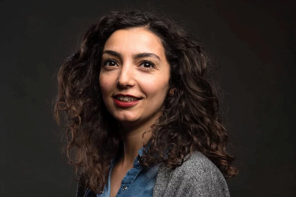 Koyu renk kıvırcık saçları, kırmızı ruju ve neşeli gülümsemesiyle dikkat çeken Janet Barış, açık mavi kot gömlek ve gri hırka giymiş bir şekilde, koyu renk sade bir arka plan önünde hafifçe yukarıya bakarak poz veriyor.