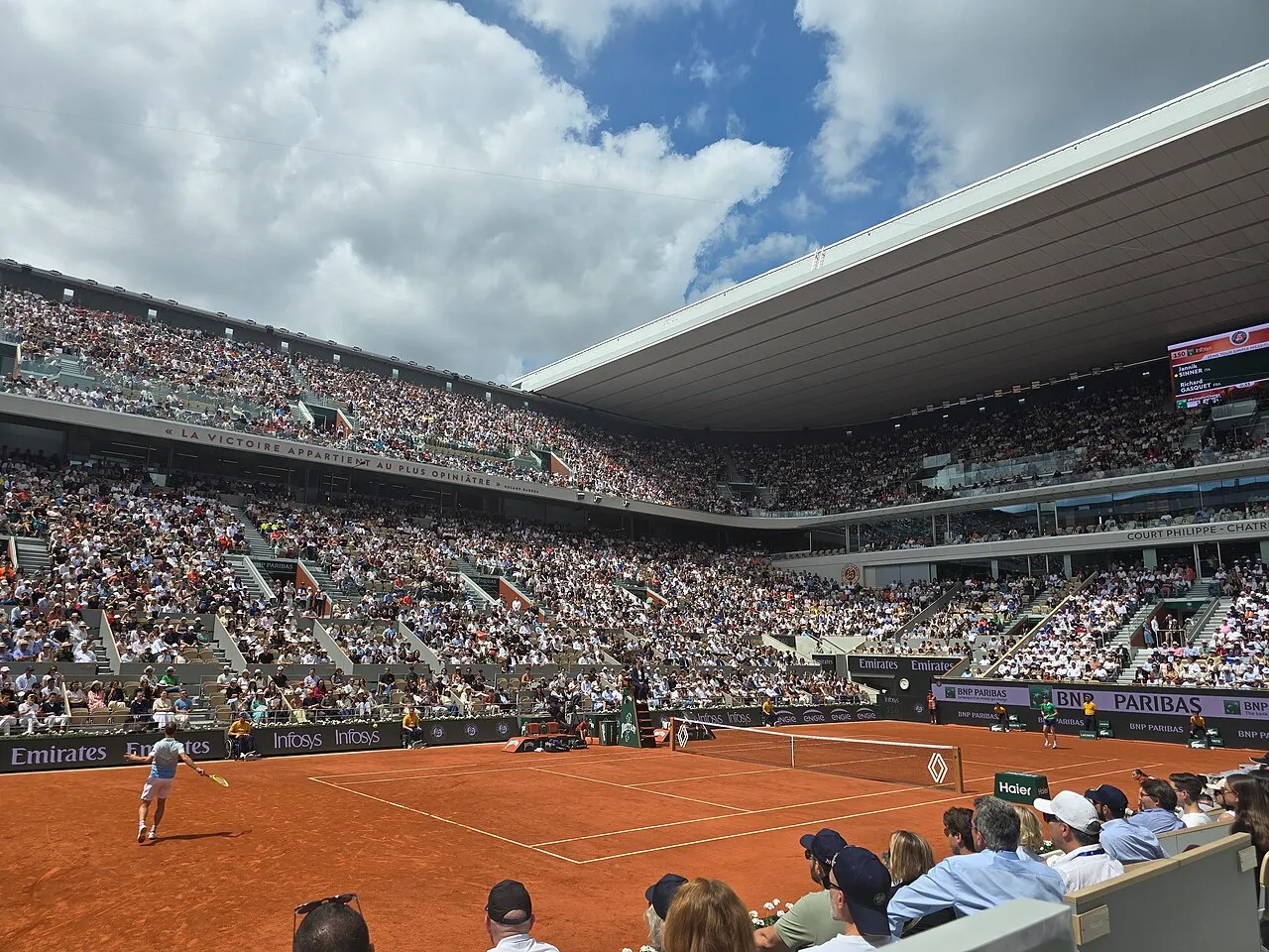 Roland Garros'taki Philippe-Chatrier kırmızı toprak kortunda Jannik Sinner ve Richard Gasquet arasında geçen tenis maçını izleyen, her bir kademesi kalabalık seyircilerle dolu tribünler ve parçalı bulutlu bir gökyüzü görülüyor.