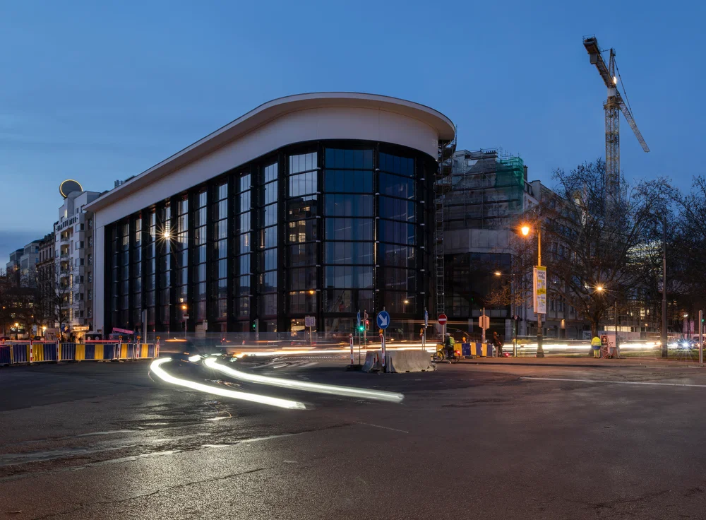 Akşam alacakaranlığında çekilen bu uzun pozlama fotoğrafında, Brüksel'deki Kanal-Centre Pompidou'nun modern, kavisli çatılı ve cam cepheli mimarisi, caddeden geçen araçların oluşturduğu parlak ışık izleriyle aydınlanıyor ve sağda bir inşaat vinci yükseliyor.
