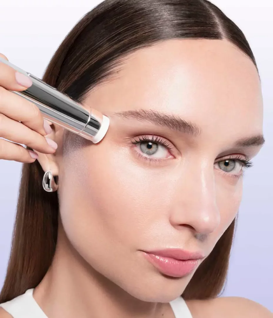 Açık renk gözlere ve ortadan ayrılmış kahverengi saçlara sahip bir kadın, sol elindeki gümüş renkli, modern bir cilt bakım cihazını alnına nazikçe uygulayarak Lancome Nano-Resurfacer 400 Booster Starter ürününü pürüzsüz ve parlak bir cilt görünümü için kullanıyor.