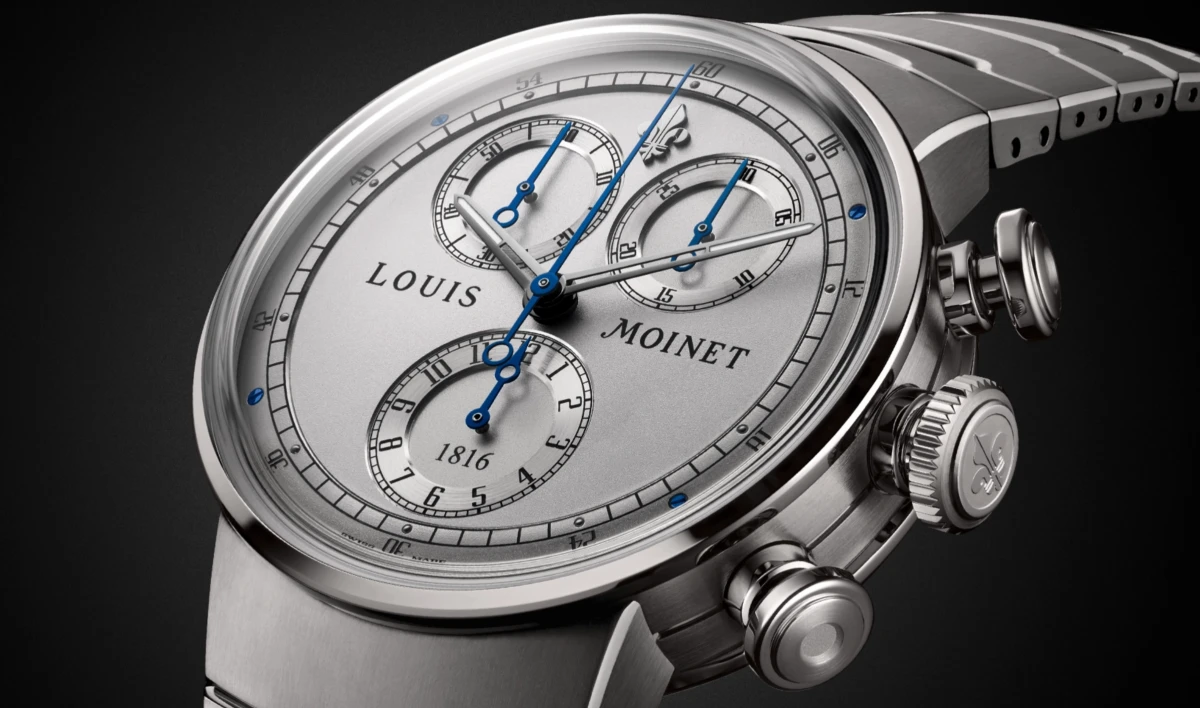 Bir Louis Moinet 1816 kronograf modelini sergileyen bu yakın çekimde, gümüş rengi şık kadran, zarif mavi ibreler, üç hassas alt kadran ve yan tarafındaki işlemeli kurma koluyla birlikte lüks ve sofistike tasarım detayları vurgulanarak, saatin işçiliğini ve estetiğini ön plana çıkarıyor.