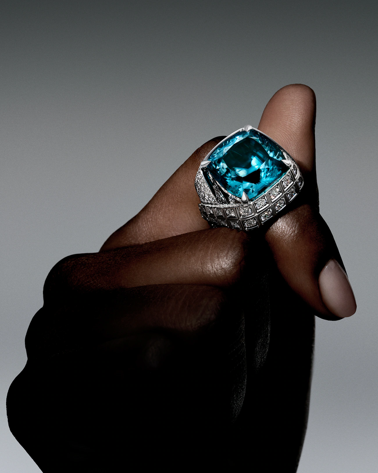 Koyu tenli bir elin başparmağında duran, Louis Vuitton Mythica Koleksiyonu’ndan Fortitude yüzük, merkezindeki büyük ve yoğun mavi cushion-step-cut zirkon taşını, beyaz altın üzerine işlenmiş ışıltılı pırlanta detaylarıyla çevreleyerek cesaret ve dayanıklılık temalarını göz alıcı bir zarafetle vurguluyor.