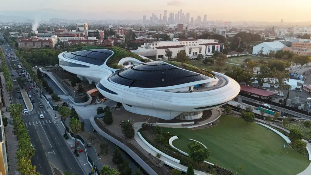 Lucas Narrative Sanat Müzesi'nin Los Angeles şehir merkezinin silüeti önünde, güneş panelleriyle süslenmiş kıvrımlı beyaz mimarisi ve yemyeşil peyzajlı çevresiyle gün batımında çekilmiş kuşbakışı görüntüsü, modern sanat ve sürdürülebilirliğin birleşimini sergiliyor.