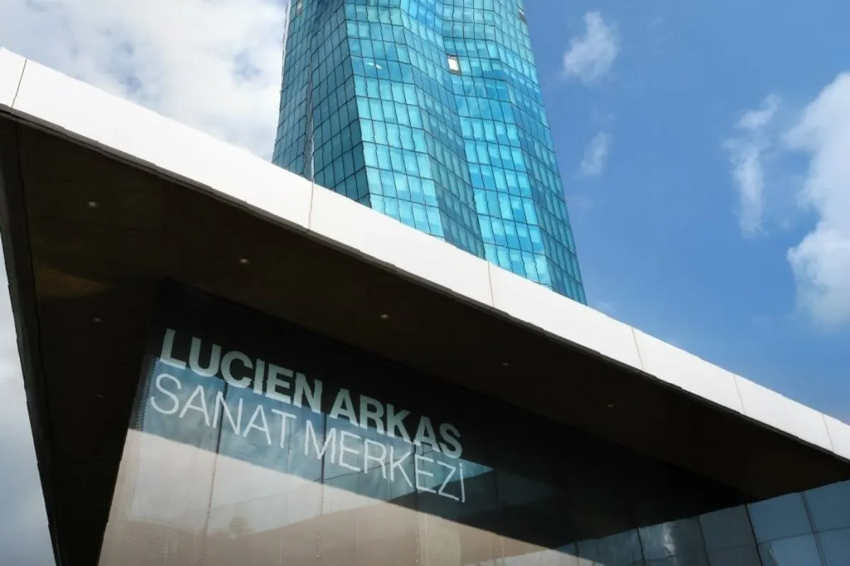 Bulutlu masmavi gökyüzünün altında yükselen, kıvrımlı hatlara sahip modern mavi cam cepheli yüksek bir binanın önünde, giriş saçağının altındaki cam yüzeyde belirgin beyaz harflerle Lucien Arkas Sanat Merkezi yazan şık mimari bir detayın alttan çekilmiş geniş açılı görüntüsü.