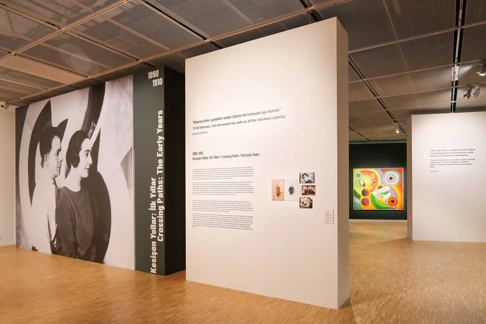 Lucien Arkas Sanat Merkezi'ndeki Kesisen Yollar: İlk Yıllar sergisinden geniş bir görünüm, solda büyük bir siyah beyaz portre ve sergi bilgisini içeren panellerle, arka planda renkli soyut bir tabloyla modern bir sanat galerisi atmosferini yansıtıyor.