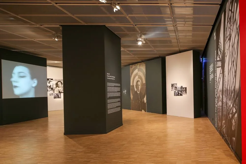 Lucien Arkas Sanat Merkezi'ndeki bu geniş ve modern sergi alanında, solda siyah beyaz bir kadın yüzü projeksiyonu, ortada hedef tahtasını andıran bir erkek portresi ve sağda ağsı bir desene sahip büyük panellerin yer aldığı, parke zeminli aydınlık bir galeri içi görünümü.