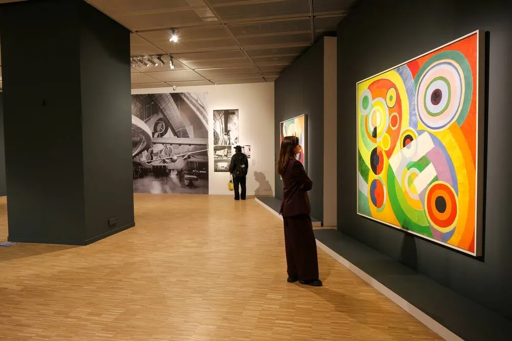 Lucien Arkas Sanat Merkezi'nin geniş sergi alanında, koyu renkli duvarlara asılı canlı tonlardaki soyut bir tabloyu büyülenmişçesine izleyen bir kadın ve ilerideki siyah beyaz fotoğraflara göz atan başka bir ziyaretçi, sanat deneyimini yansıtıyor.