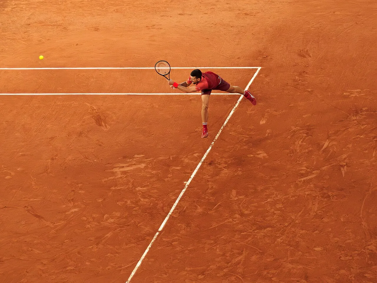 Roland-Garros'ta toprak kort üzerinde çekilmiş yukarıdan bir görüntüde, kırmızı tişört ve şort giyen Novak Djokovic, sarı bir tenis topuna sağ elindeki raketiyle uzanarak çevik bir şekilde yere doğru eğilirken, top kort çizgilerinin yanında havada süzülüyor.