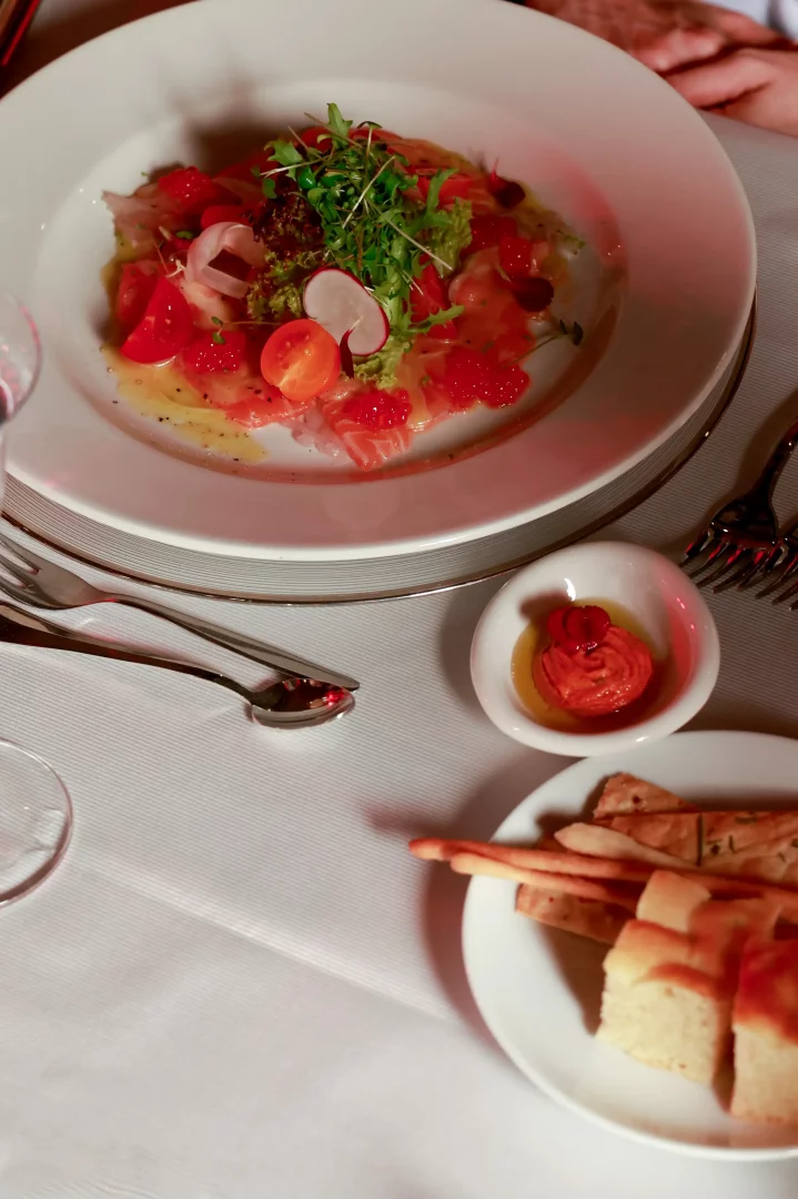 Beyaz masa örtüsü üzerinde, somon carpaccio, çeri domatesler, parlak somon havyarı, taze yeşillikler ve ince turp dilimleriyle özenle hazırlanmış şık bir başlangıç tabağı, Frederic Malle menüsünün görsel şölenini yansıtırken, yanında küçük bir sosluk ve çeşitli ekmekler de masada yer alıyor.