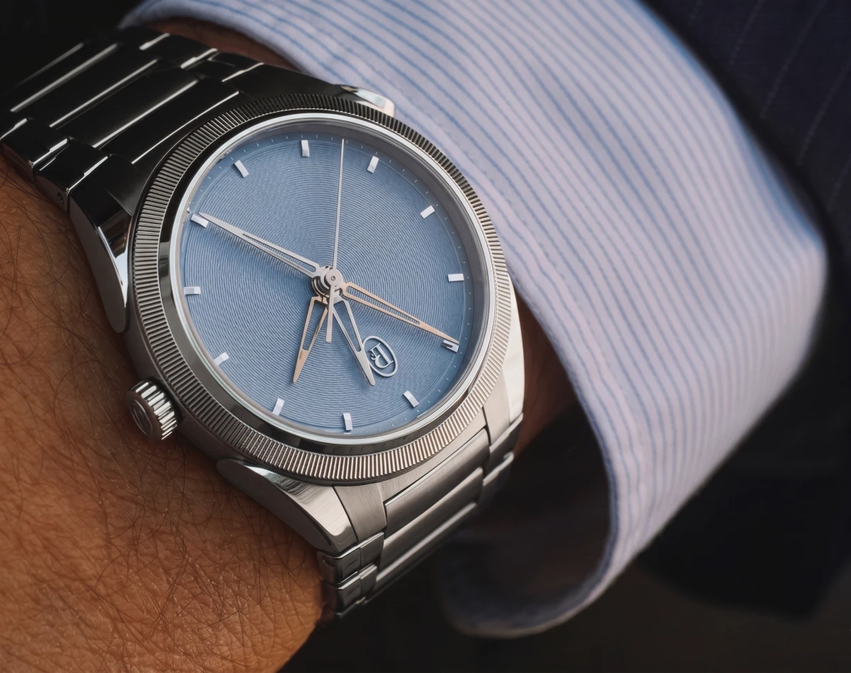 Parmigiani Fleurier Tonda PF Chronographe Mysterieux kronograf saati, zarif çizgili gömlek manşeti ve koyu renk bir takım elbise detayıyla çevrili bir bilekte, dairesel desenli açık mavi kadrana ve parlak metal bileziğe sahip.