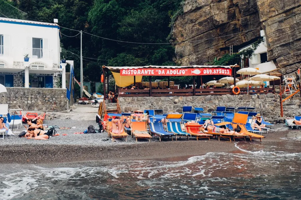 Çakıllı Positano sahilinde, dalgaların kıyıya vurduğu denizin kenarında, renkli şezlonglarda uzanan ve Ristorante Da Adolfo adlı restoranın altındaki masalarda yemek yiyen insanların olduğu, ardında yeşil bitki örtüsüyle kaplı yüksek kayalıkların yükseldiği canlı bir İtalya plaj manzarası.