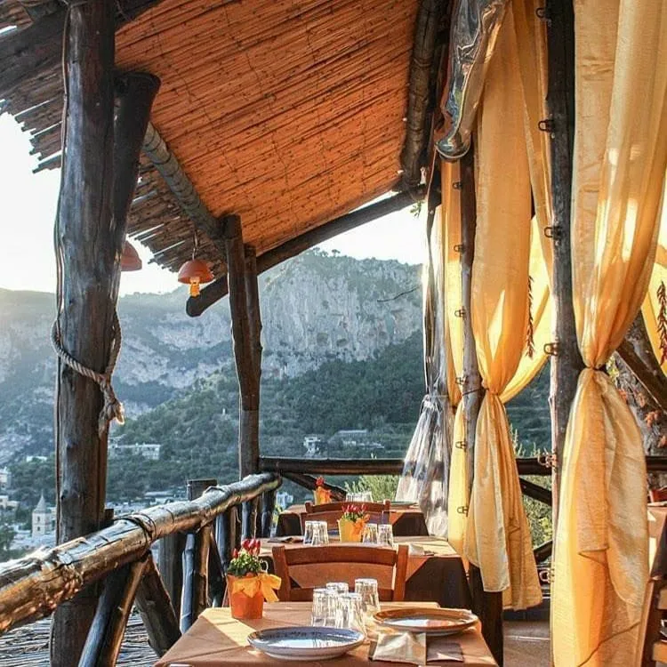 Dağlık bir vadiye bakan, ahşap kolonlarla desteklenmiş ve hasır tavanlı bir açık hava restoranının terasında, gün batımının sıcak ışığı altında hazır bekleyen kahverengi masa örtülü masalar, zarif sarı perdeler ve uzakta görünen evler büyüleyici bir manzara sunuyor.