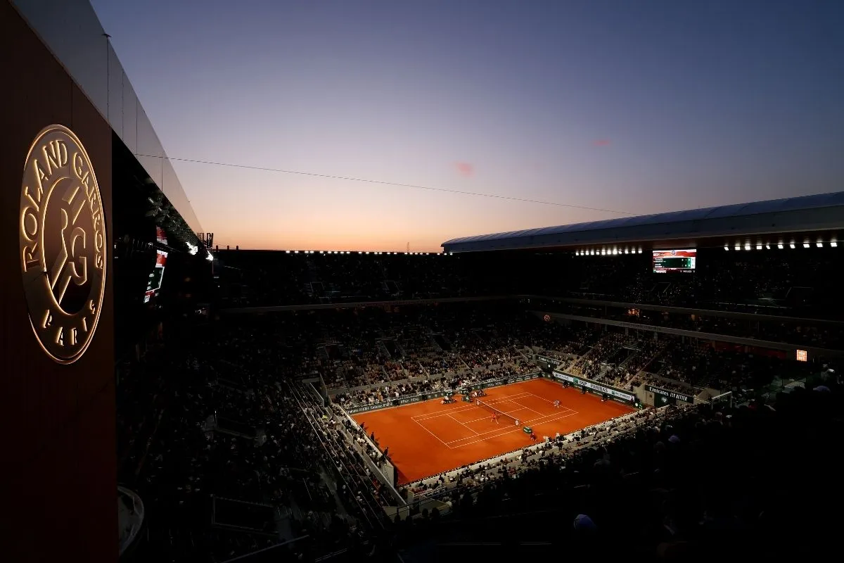 Gün batımında aydınlanan kırmızı toprak kortta devam eden bir Roland Garros (Fransa Açık) maçını kuşbakışı gösteren bu geniş açılı görselde, tribünlerdeki kalabalık siluetler ve sol üst köşede parlayan turnuva logosu, 2026 takvimi detaylarını içeren bu rehberdeki turnuvanın eşsiz atmosferini vurguluyor.