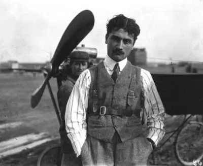 1910'lu yılların başlarında çekilmiş bu siyah beyaz fotoğrafta, koyu bıyıklı ve saçlı, çizgili gömlek, kravat ve askılı şık bir yelek giyen havacı Roland Garros, elleri cebinde kameraya ciddi bir ifadeyle bakarken, arkasında büyük ahşap pervaneli erken dönem bir uçak ve kısmen görünen bir görevli yer alıyor.