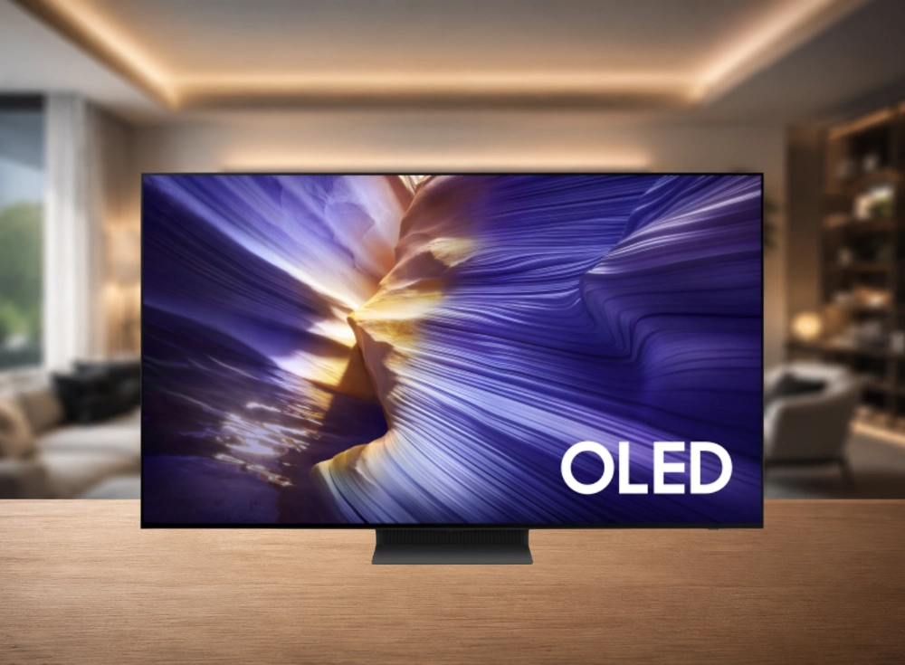 Ahşap bir masa üzerinde duran şık ve ince çerçeveli Samsung OLED televizyon, modern bir oturma odasının ambiyansında, ekranında mor, mavi ve sarı tonlarda soyut bir görselle ve sağ alt köşesinde belirgin OLED yazısıyla üstün bir oyun televizyonu deneyimi vadediyor.
