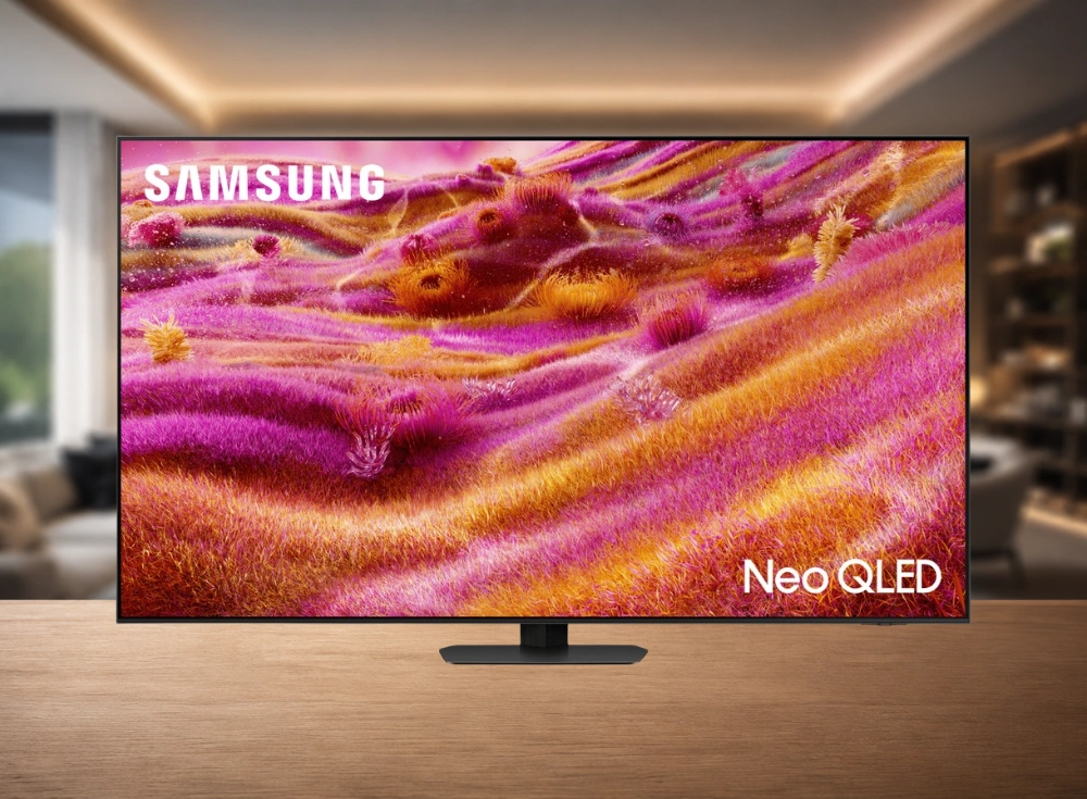 Arka planı bulanık, modern bir oturma odasında ahşap bir masa üzerinde duran, ekranında pembe, mor, turuncu ve sarı tonlarda dalgalı, canlı ve soyut bir manzara gösteren 65 inç Samsung Neo QLED Mini LED 4K akıllı televizyon.