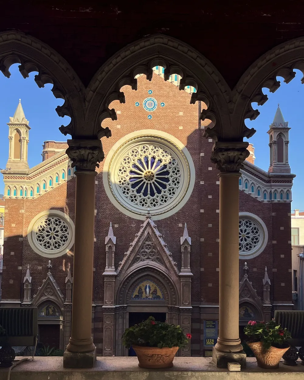 Sant'Antonio Kilisesi'nin detaylı mimarisi, tuğla cephesi üzerindeki büyük Gotik gül pencereleri ve iki zarif çan kulesiyle, oymalı taş kemerli bir avlunun içinden masmavi gökyüzüne doğru uzanırken, ön planda yer alan saksı çiçekleri bu etkileyici yapıyı zarifçe çerçeveliyor.