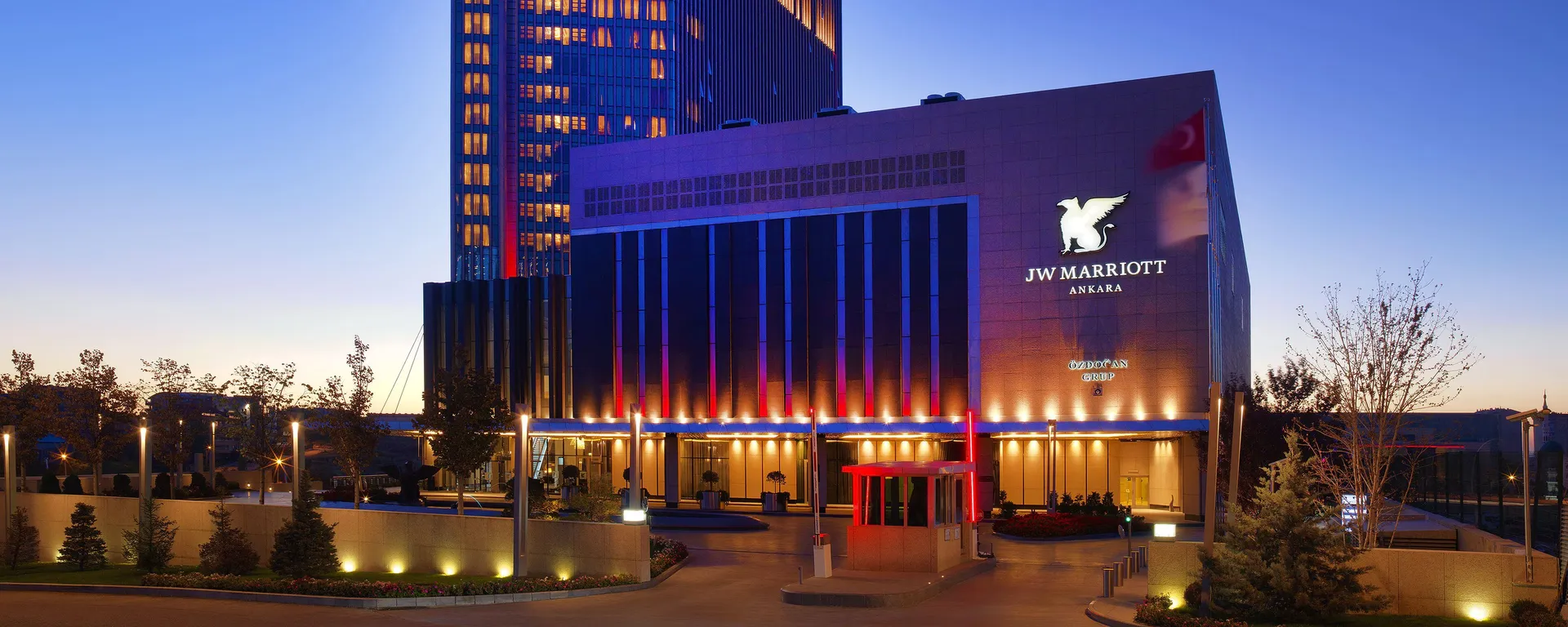 JW Marriott Ankara otelinin modern mimarisi, gün batımından geceye geçişin eşsiz anında gökyüzünün mavi ve turuncu tonları altında, cephesindeki kırmızı ve mavi vurgu ışıklarıyla aydınlanarak büyüleyici bir görüntü sergiliyor.