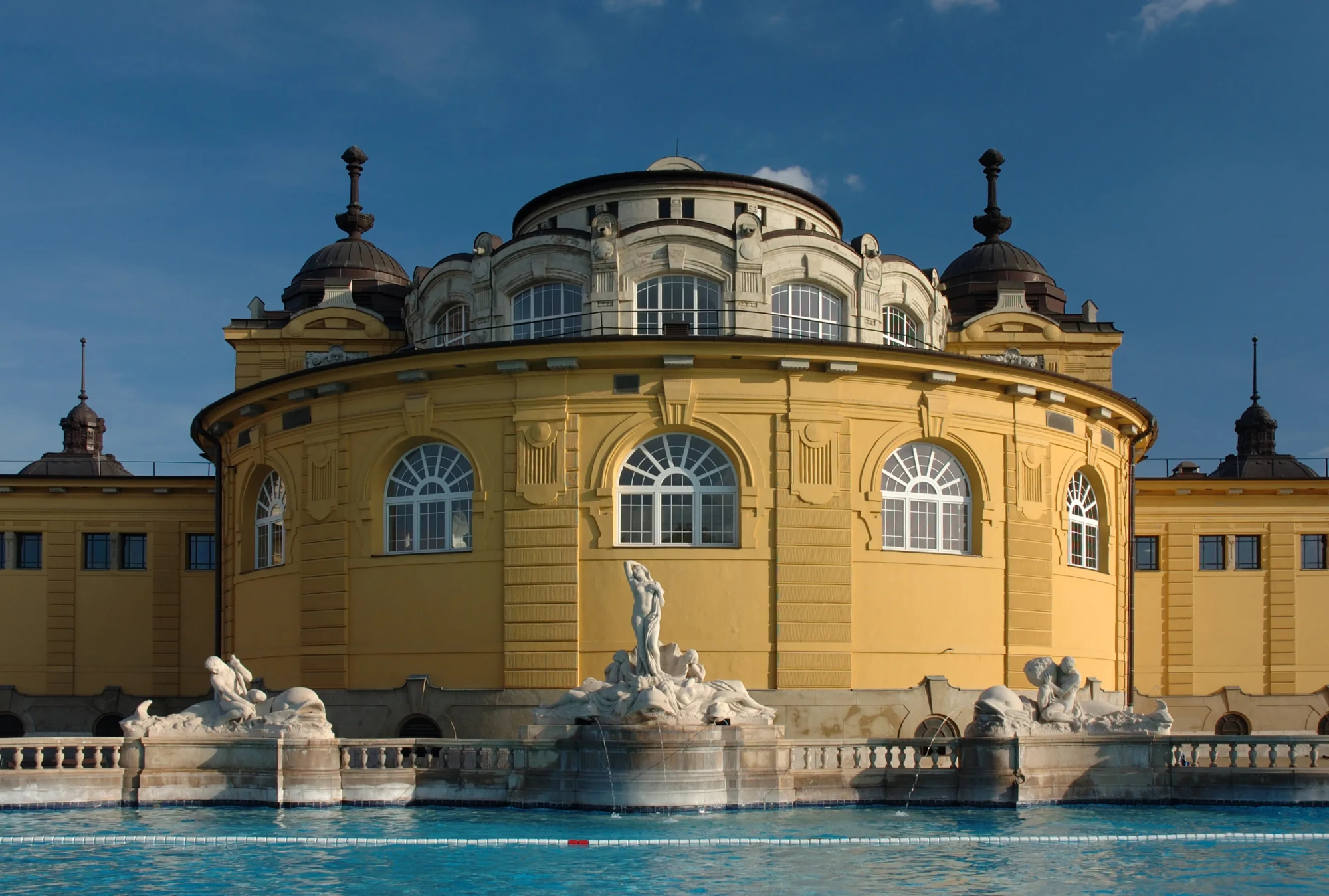 Budapeşte'deki Széchenyi Termal Hamamı'nın dış cephesini gösteren bu fotoğraf, kubbelerle süslü, sarı renkli, görkemli binanın önündeki berrak mavi havuzda fışkıran heykelli çeşmeleri ve zarif mimarisini güneşli bir günde gözler önüne seriyor.