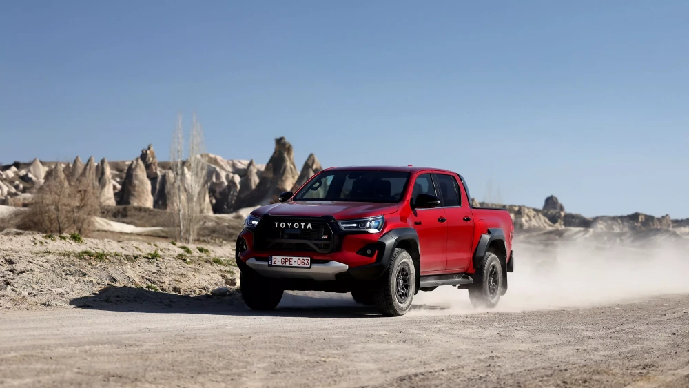 Kırmızı bir Toyota Hilux GR SPORT pickup, kumlu ve engebeli bir arazi yolunda ilerlerken, arka planda belirgin kaya oluşumları ve kuru ağaçlar seçiliyor, tekerleklerinden yükselen tozlar aracın dinamik hareketini vurguluyor.