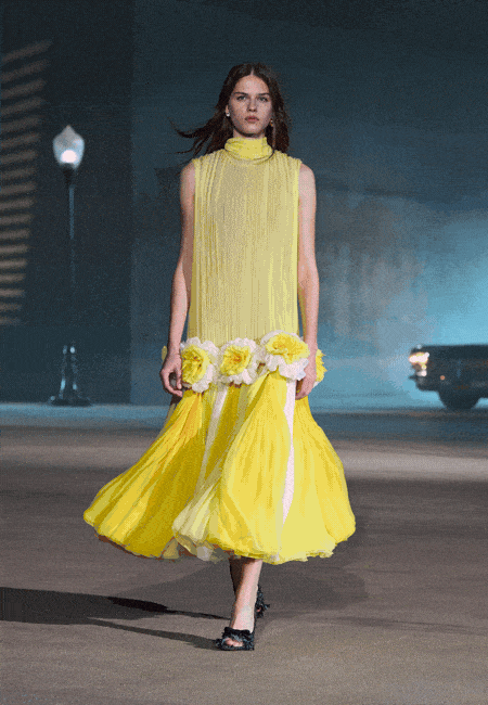Jonathan Anderson'ın Dior Cruise 2027 koleksiyonundan bir model, Hollywood cazibesini ve California ruhunu yansıtan, belinde iri beyaz-sarı California poppy çiçekleri ve eteklerinde beyaz şerit detayları olan, uçuş uçuş parlak sarı, yüksek yakalı, pileli kolsuz bir elbiseyle loş ışıklı podyumda zarifçe ilerliyor.