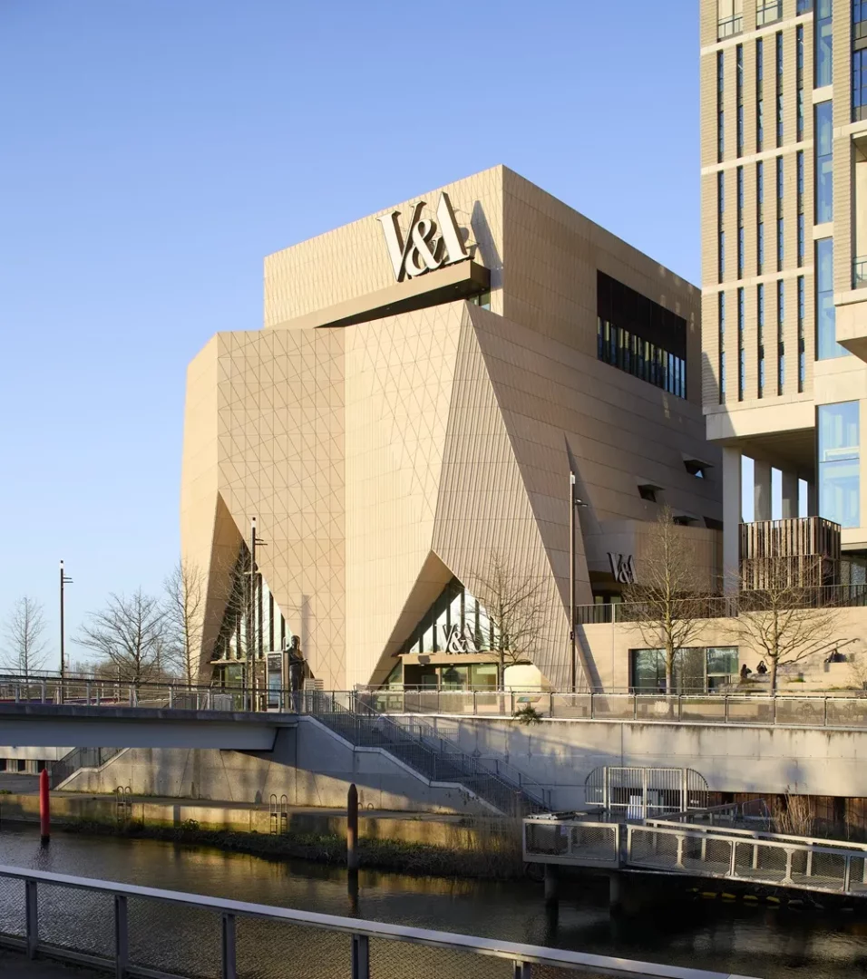 V&A East Müzesi'nin kendine özgü, geometrik ve açılı bej cephesiyle öne çıkan modern binası, heykelli bir köprünün ve su yolunun kıyısında, yanındaki uzun yapıyla birlikte açık mavi gökyüzünün altında çarpıcı bir şekilde konumlanıyor.
