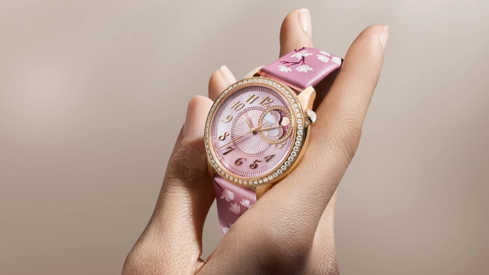 Pembe altın kasalı, pırlanta çerçeveli ve pembe sedef kadranlı Vacheron Constantin Egerie Moon Phase Spring Blossom sınırlı üretim saatin, beyaz çiçek desenli pembe kayışı ve ay evresi göstergesi yakın çekimde bir el üzerinde zarifçe duruyor.