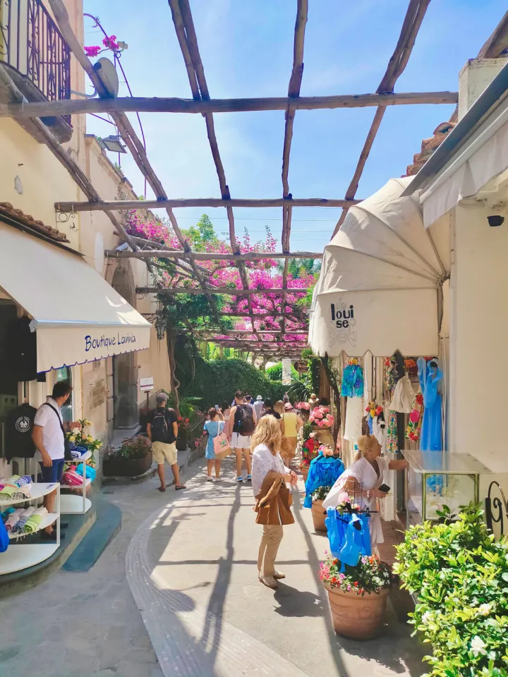 Dar, taş döşemeli bir sokakta, ahşap pergolanın pembe begonvillerle kaplandığı gölgelerin altında, çeşitli butiklerin ve dükkanların önünden geçen kalabalık turistler, sıcak bir Akdeniz kasabasının canlı ve hareketli atmosferini yansıtıyor.