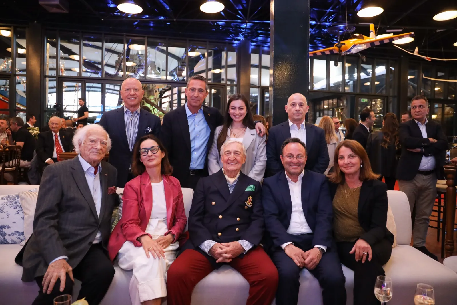 Şık bir iç mekanda, tavandan sarkan model uçaklar ve büyük cam pencerelerle çevrili bir etkinlikte, ön planda oturan beyaz saçlı yaşlı bir beyefendi, pembe blazer ceketli bir kadın ve lacivert kruvaze ceketli başka bir beyefendinin de dahil olduğu, takım elbiseli ve zarif kıyafetli on bir kişilik bir grup fotoğraf için poz veriyor.