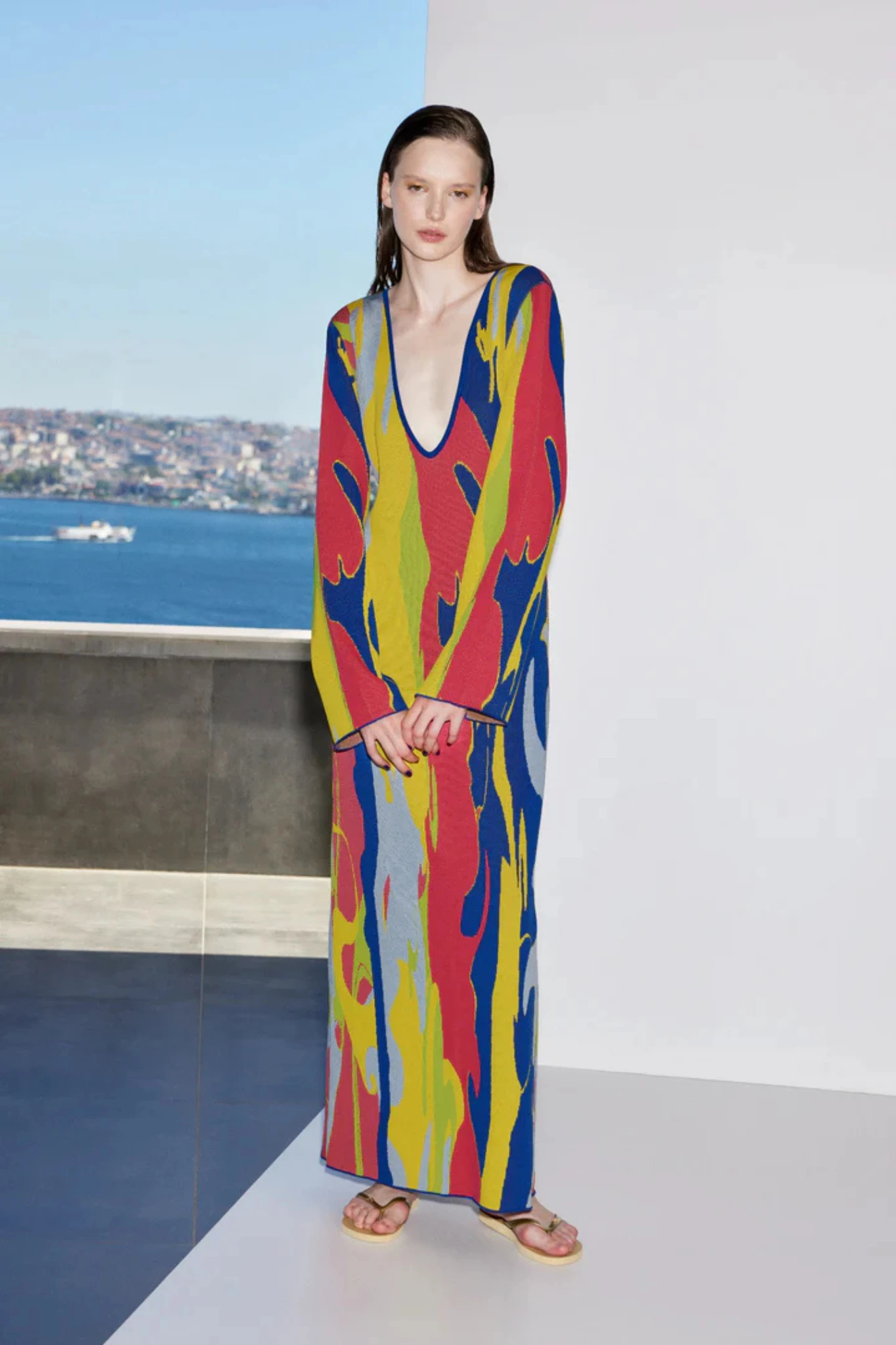 Model, Ace Nayman'ın Sunnylands koleksiyonundan, Mark Rothko'dan ilham alan canlı kırmızı, sarı ve mavi tonlarında AI destekli soyut desenlere sahip, derin V yaka uzun triko elbisesi ve altın renkli terlikleriyle, modern kadının enerjisini yansıtan şehir ve deniz manzaralı bir stüdyoda duruyor.