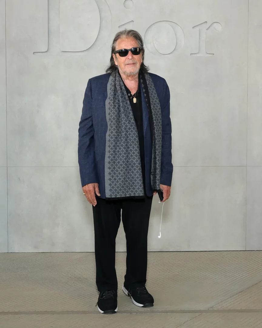 Al Pacino, Jonathan Anderson'ın Los Angeles'ta düzenlediği ilk Dior Cruise 2027 koleksiyonu defilesinde, gri Dior logosu önünde lacivert blazer ceketi, desenli gri atkısı, siyah güneş gözlükleri ve spor ayakkabılarıyla Hollywood ihtişamını yansıtan bir duruş sergiliyor.