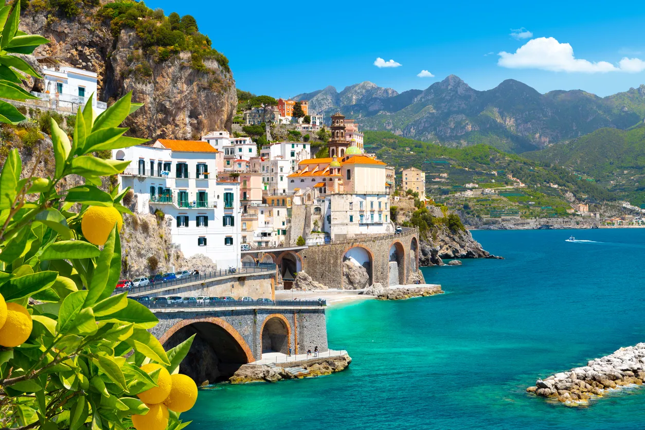 Amalfi Sahili'nin masmavi suları ve dik kayalıklarına inşa edilmiş, beyaz evleri ve tarihi kilisesiyle ünlü büyüleyici kasabası, ön plandaki yeşil yapraklı ve sarı limonlu dallarla çerçevelenerek, bu gezi rehberi için eşsiz bir görünüm sunuyor.